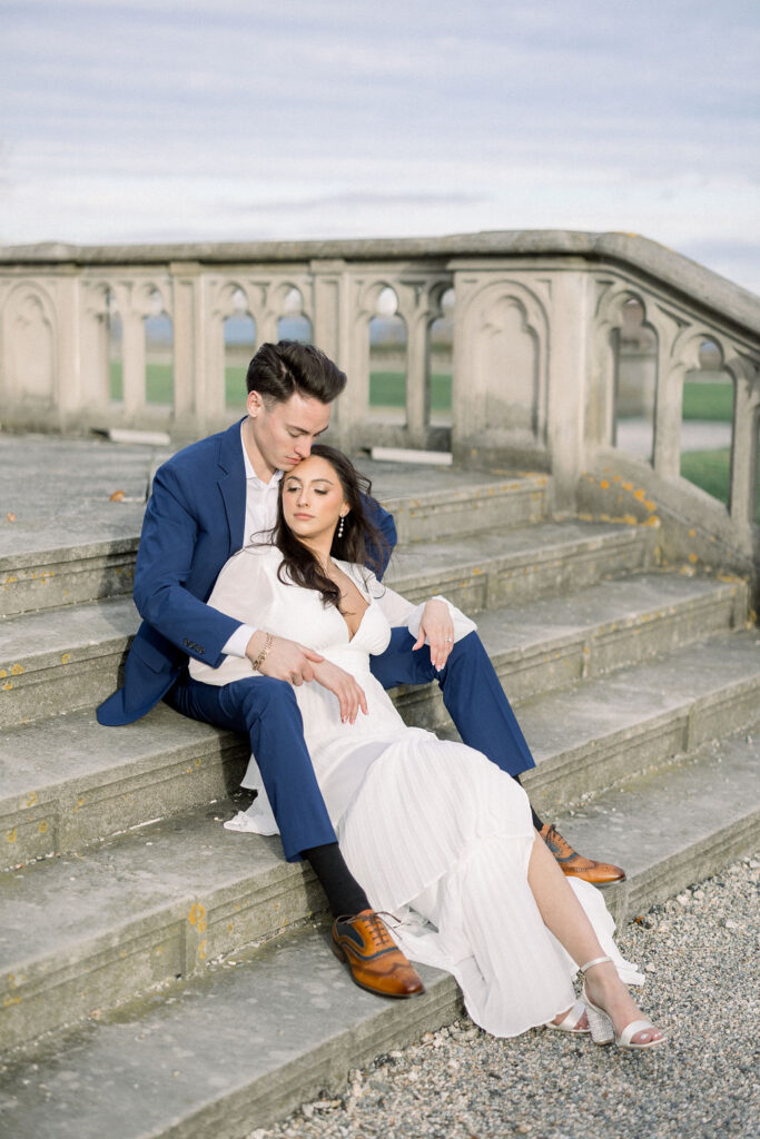 Newport Beach Wedding Venues | Brianna Graca Weddings Fine Art Wedding Photographer in Newport | A couple sits on stone steps; the man in a blue suit embraces the woman in a white dress, conveying tenderness against an ornate balustrade backdrop.