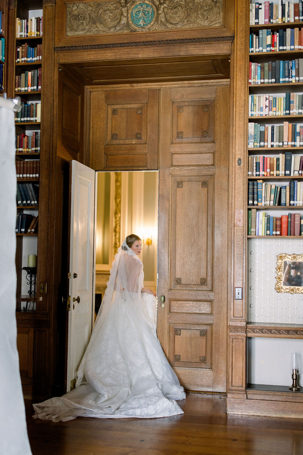 Aldrich Mansion RI | Brianna Graca Weddings Fine Art Wedding Photographer in Newport | Bride in a white gown walking through elegant wooden library doors, surrounded by bookshelves. She looks back with a serene and joyful expression.