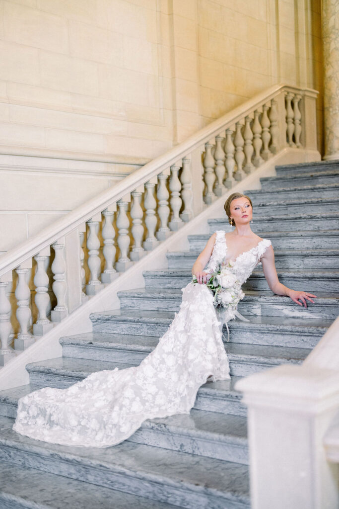 Aldrich Mansion RI | Brianna Graca Weddings Fine Art Wedding Photographer in Newport | Bride in a flowing white lace gown sits elegantly on a marble staircase, holding a bouquet of white flowers. The setting is grand and elegant.