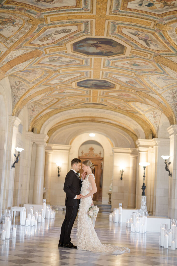 Aldrich Mansion RI | Brianna Graca Weddings Fine Art Wedding Photographer in Newport | Bride and groom embrace under an ornate, arched ceiling with intricate gold details. The elegant hallway is lined with lit candles, creating a romantic atmosphere.