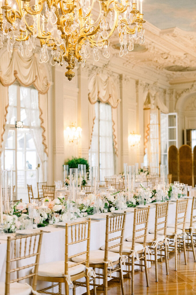 Rosecliff Mansion Wedding Photos | Brianna Graca Weddings Fine Art Wedding Photographer in Newport | Elegant dining room with a long table set for an event, adorned with white and pastel floral arrangements and gold chairs. A large ornate chandelier hangs overhead.