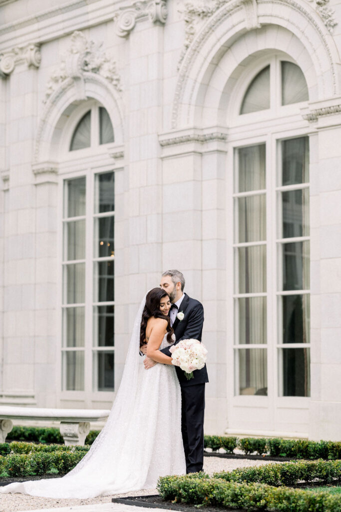 Wedding Photography RI | Brianna Graca Weddings Fine Art Wedding Photographer in Newport | A bride and groom embrace outside an elegant building with arched windows. He kisses her forehead, she holds a bouquet of white flowers. Romantic tone.