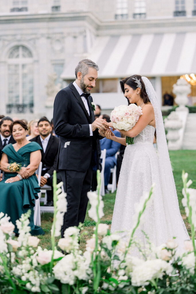 Rosecliff Mansion Wedding Photos | Brianna Graca Weddings Fine Art Wedding Photographer in Newport | A bride and groom exchange rings outdoors, surrounded by guests. The bride holds a bouquet of pale roses, wearing a sparkling gown and veil. Elegant and joyful.