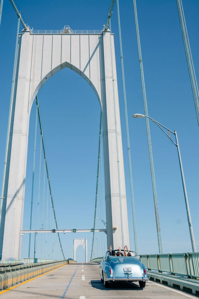 Newport Beach Wedding Venues | Brianna Graca Weddings Fine Art Wedding Photographer in Newport | A light blue convertible car drives across a suspension bridge under a clear blue sky. Passengers have their arms raised, suggesting excitement and freedom.