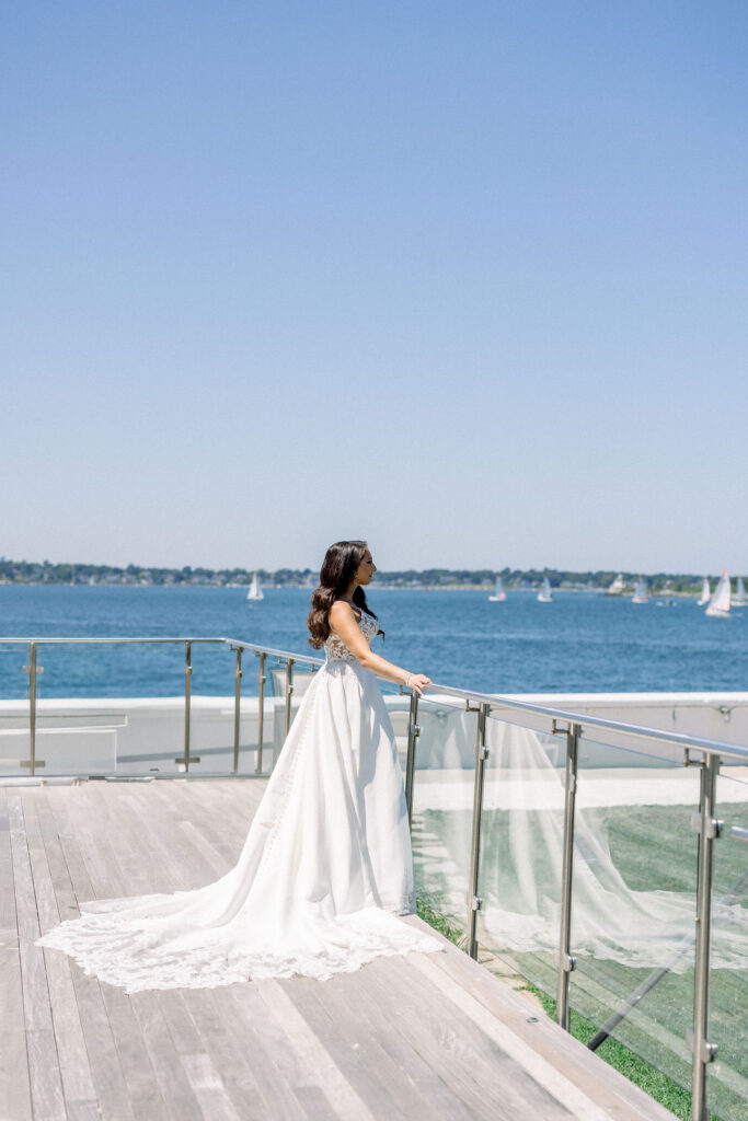 Newport Beach Wedding Venues | Brianna Graca Weddings Fine Art Wedding Photographer in Newport | Bride in flowing white gown gazes out over a serene blue sea dotted with sailboats. Clear sky and modern glass terrace enhance the tranquil scene.