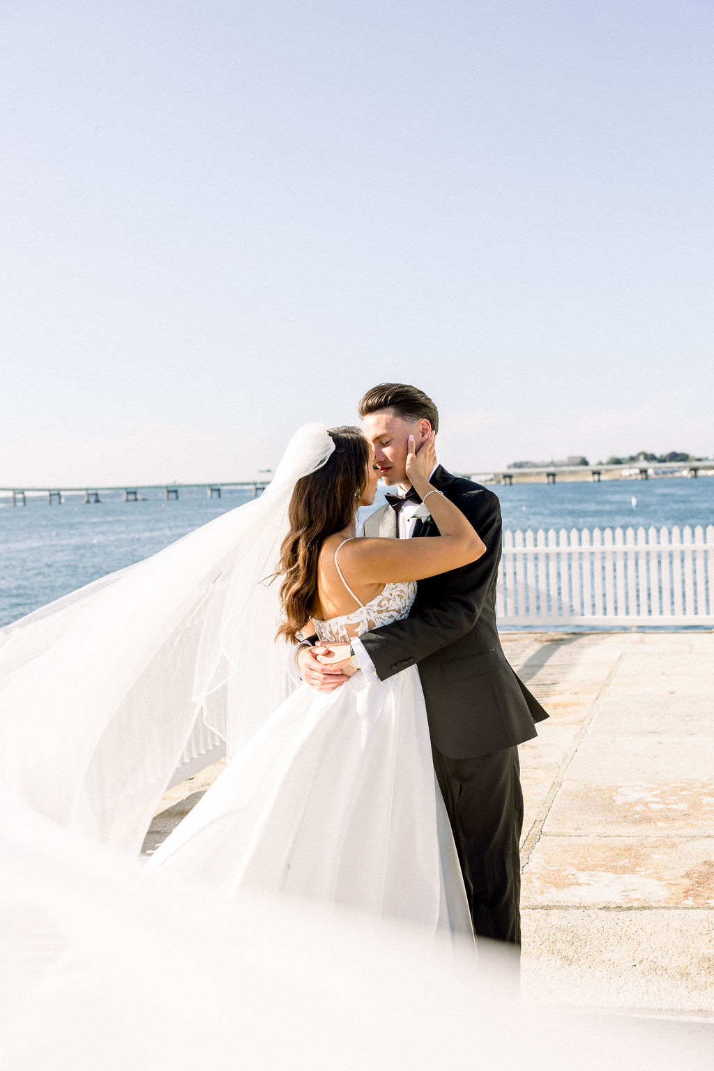 Rhode Island Wedding Photographer | Brianna Graca Weddings Fine Art Wedding Photographer in Newport | A bride in a flowing white gown and veil embraces a groom in a black suit by a waterfront, under clear blue skies, evoking joy and romance.