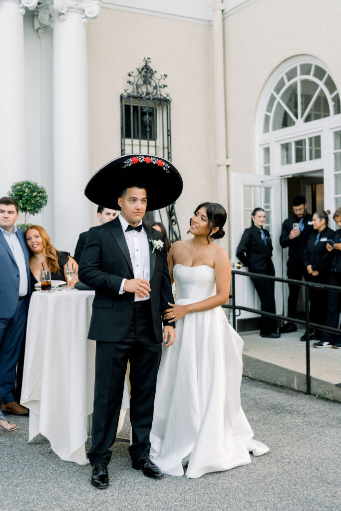 Newport Wedding Venues | Brianna Graca Weddings Fine Art Wedding Photographer in Newport | A bride in a strapless white gown smiles at a groom wearing a black tuxedo and large sombrero. They stand in front of a venue, suggesting a joyful wedding reception.