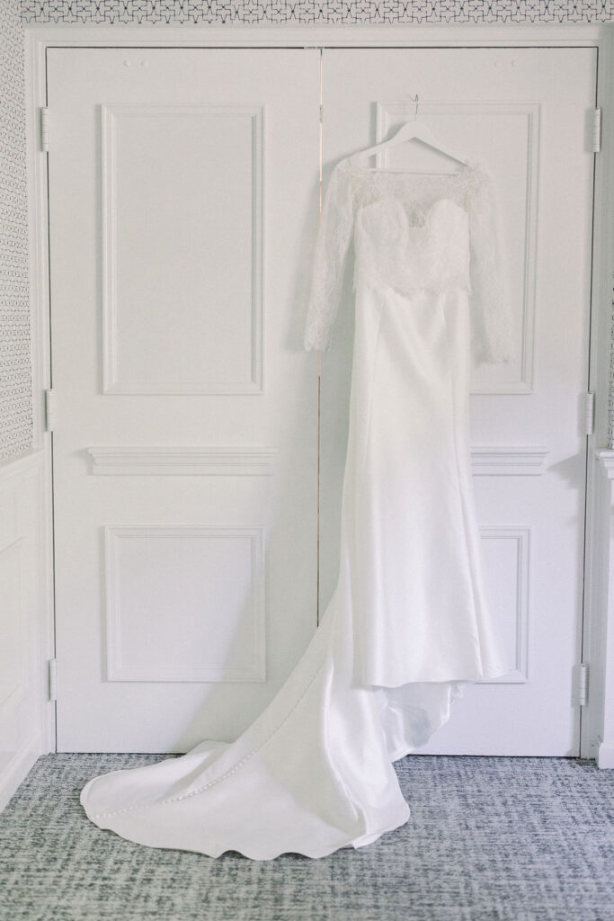 Newport Beach Wedding Venues | Brianna Graca Weddings Fine Art Wedding Photographer in Newport | Elegant white wedding dress with lace sleeves and a long train, hanging on a double door. The scene is serene and minimalistic, evoking a sense of romance.