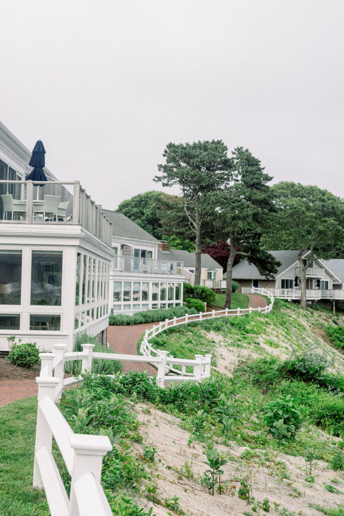 Wequassett Wedding | Brianna Graca Weddings Fine Art Wedding Photographer in Newport | Charming seaside cottages with white fences, green gardens, and brick paths under a cloudy sky. Coastal tranquility and inviting architecture.