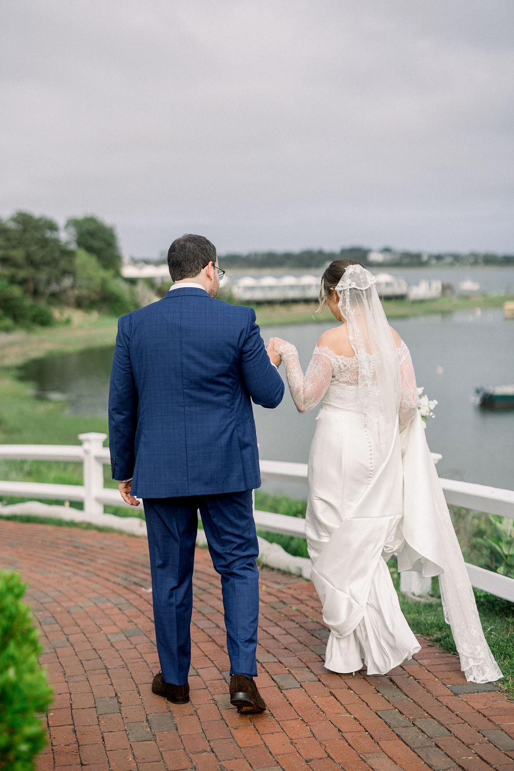 Newport Beach Wedding Venues | Brianna Graca Weddings Fine Art Wedding Photographer in Newport | A bride and groom walk hand in hand on a brick path near a scenic lake. The bride wears a white gown with lace, and the groom is in a blue suit.