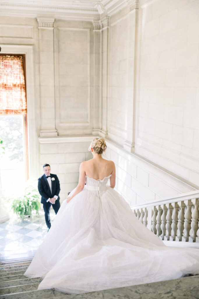 Aldrich Mansion | Newport RI Wedding Photographer Brianna Graca Weddings | Bride in elegant white gown descends marble staircase, approaching groom in a black tuxedo. Sunlit, classic interior creates a romantic atmosphere.
