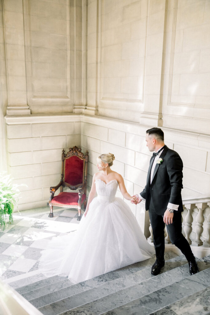 Aldrich Mansion Wedding | Brianna Graca Weddings Fine Art Wedding Photographer in Newport | Elegant couple in wedding attire on marble stairs. The bride in a flowing white gown holds hands with the groom in a black tuxedo. Red velvet chair nearby.