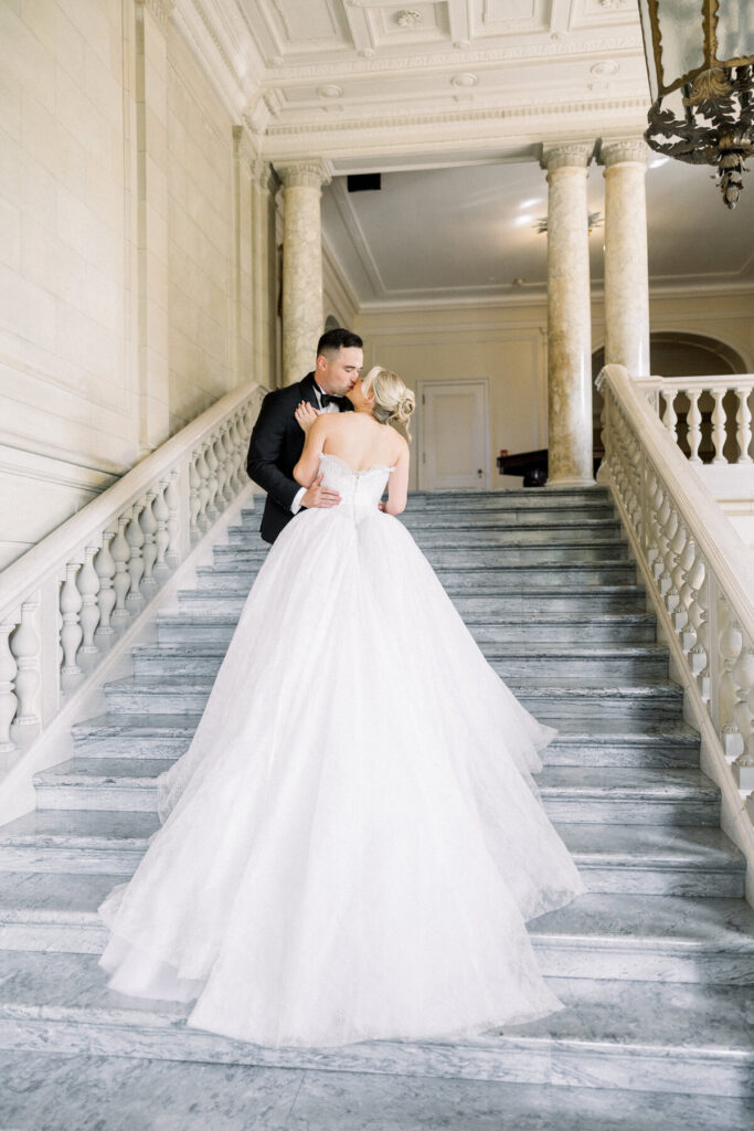 Aldrich Mansion Wedding | Brianna Graca Weddings Fine Art Wedding Photographer in Newport | A bride in a flowing white gown and a groom in a black suit embrace on elegant marble stairs in a grand hallway, conveying romance and elegance.