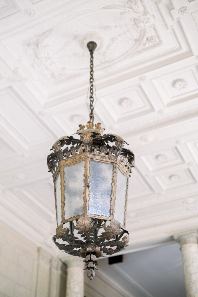 Aldrich Mansion Wedding | Brianna Graca Weddings Fine Art Wedding Photographer in Newport | Ornate vintage lantern with frosted glass panels and intricate metalwork hangs from a chain on a decorative coffered ceiling, evoking elegance.