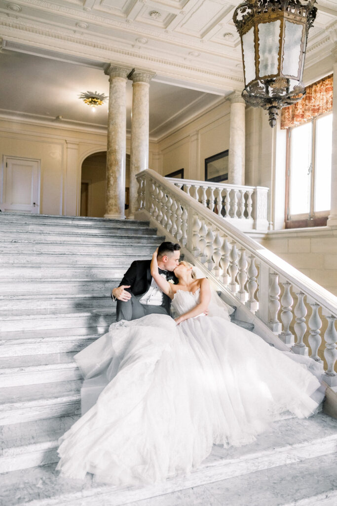 Aldrich Mansion | Newport RI Wedding Photographer Brianna Graca Weddings | A couple in wedding attire lovingly kiss while seated on elegant marble stairs. Sunlight pours through a window, highlighting their joyful moment.