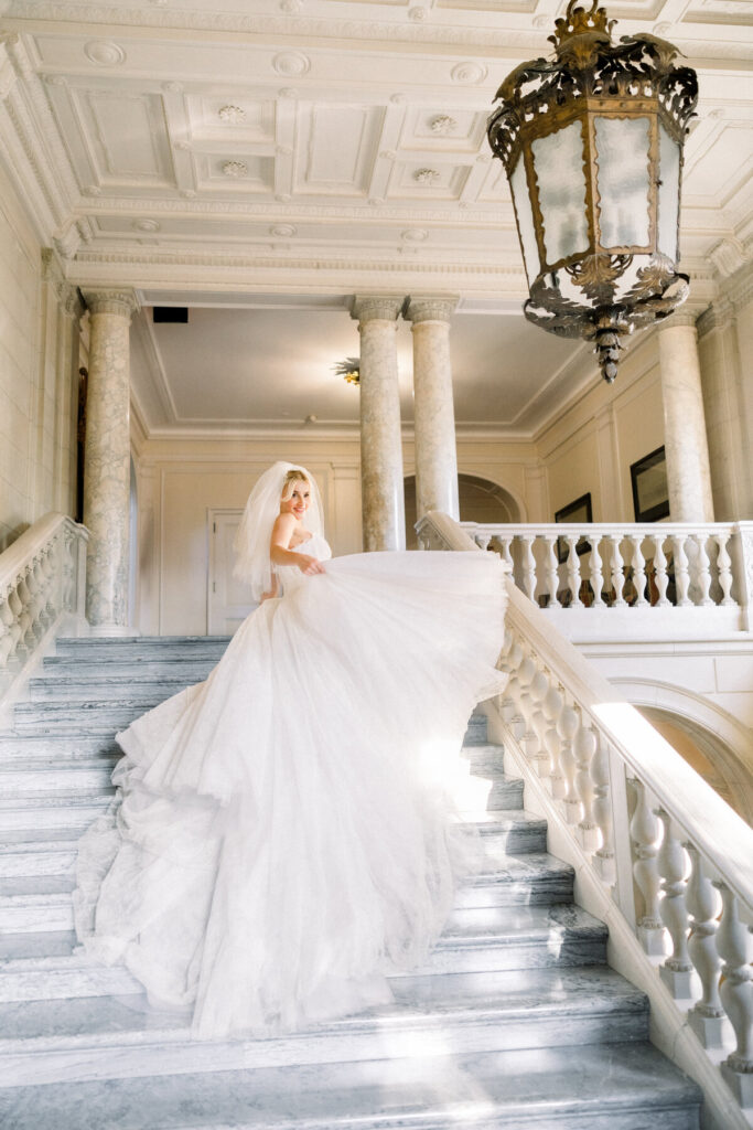 Aldrich Mansion Wedding | Brianna Graca Weddings Fine Art Wedding Photographer in Newport | A bride in a flowing white wedding gown stands on an elegant marble staircase, under soft, natural light in a grand interior with columns. Romantic and serene.
