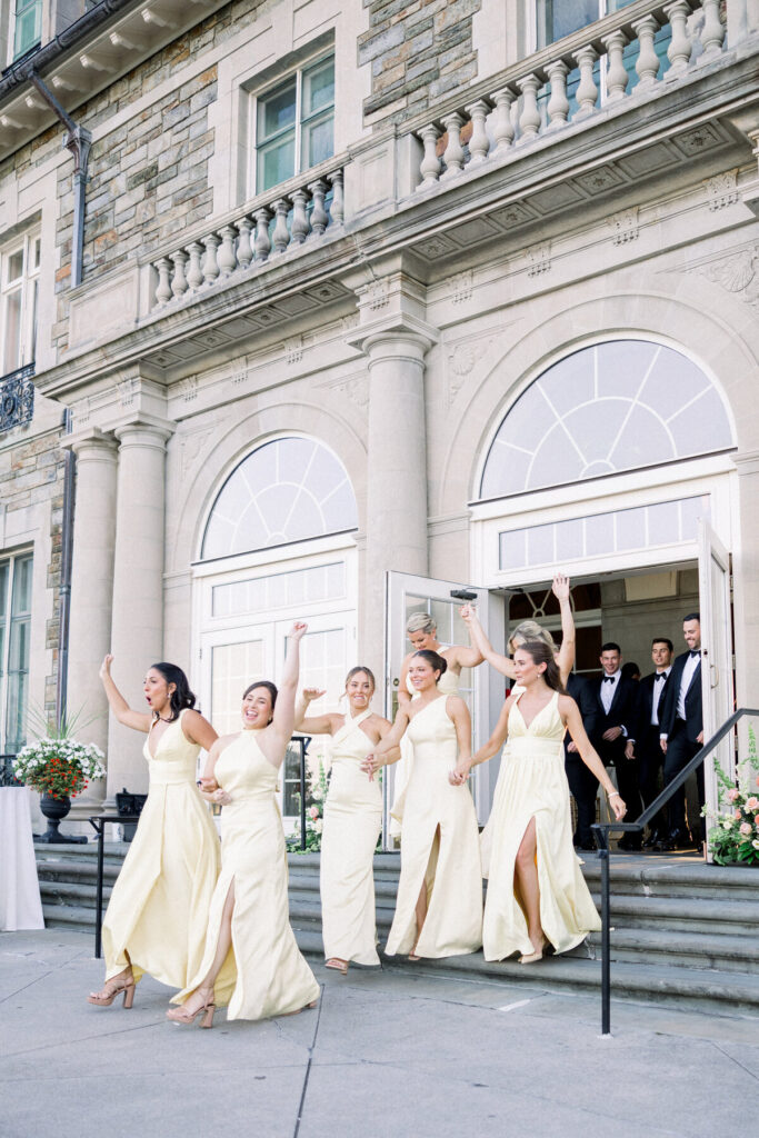 Aldrich Mansion | Newport RI Wedding Photographer Brianna Graca Weddings | A group of six bridesmaids in yellow dresses joyfully exit a building with arched windows, waving and smiling. The scene is festive and celebratory.