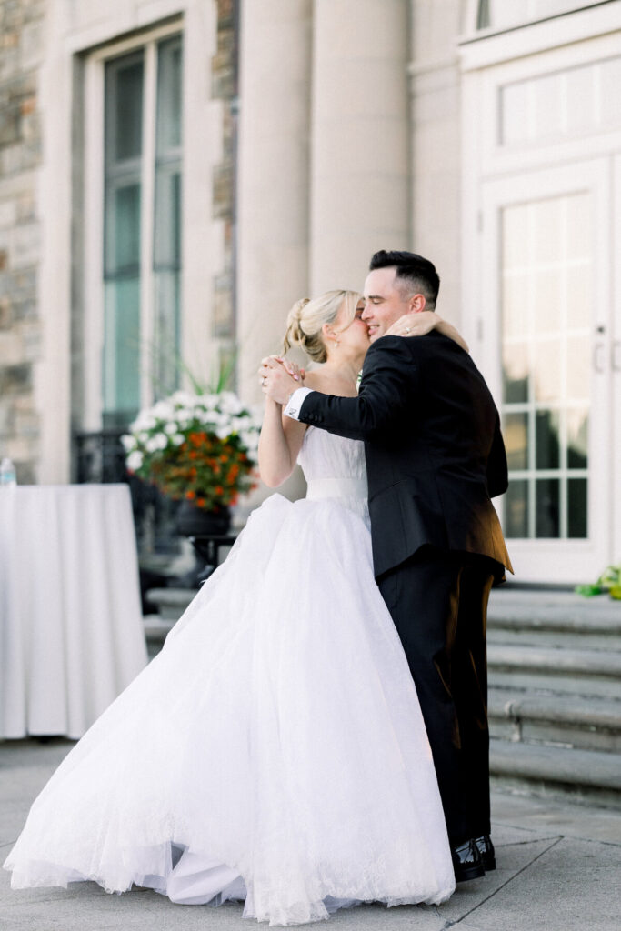 Aldrich Mansion | Newport RI Wedding Photographer Brianna Graca Weddings | A bride in a flowing white gown kisses the groom, dressed in a black suit, while dancing outside a stone building. Their embrace exudes joy and romance.
