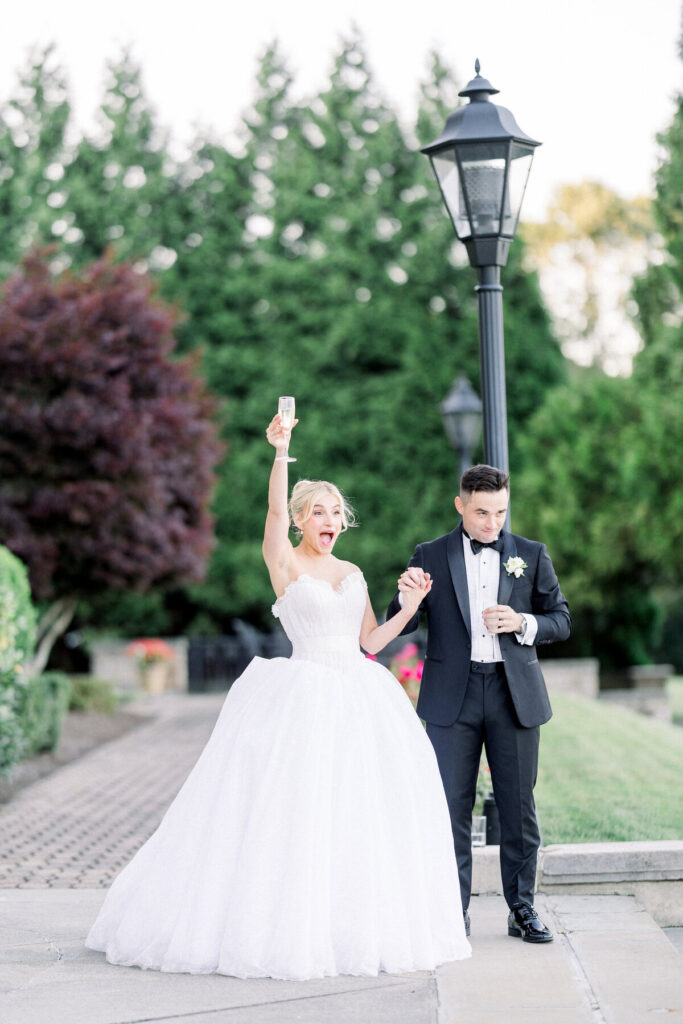 Aldrich Mansion | Newport RI Wedding Photographer Brianna Graca Weddings | Bride in a white gown joyfully raises a champagne glass next to a groom in a tuxedo. They stand on a patio with lush, green trees behind.