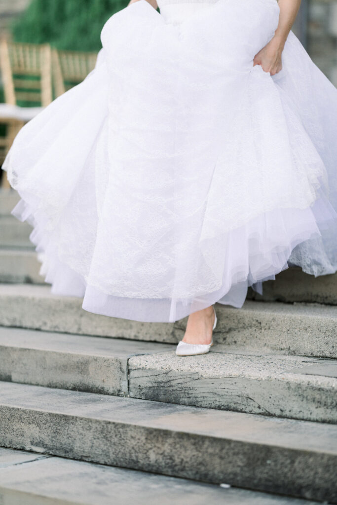 Aldrich Mansion | Newport RI Wedding Photographer Brianna Graca Weddings | A bride in a flowing white gown elegantly walks down stone steps, showcasing her pointed white shoes. The scene conveys a sense of grace and anticipation.
