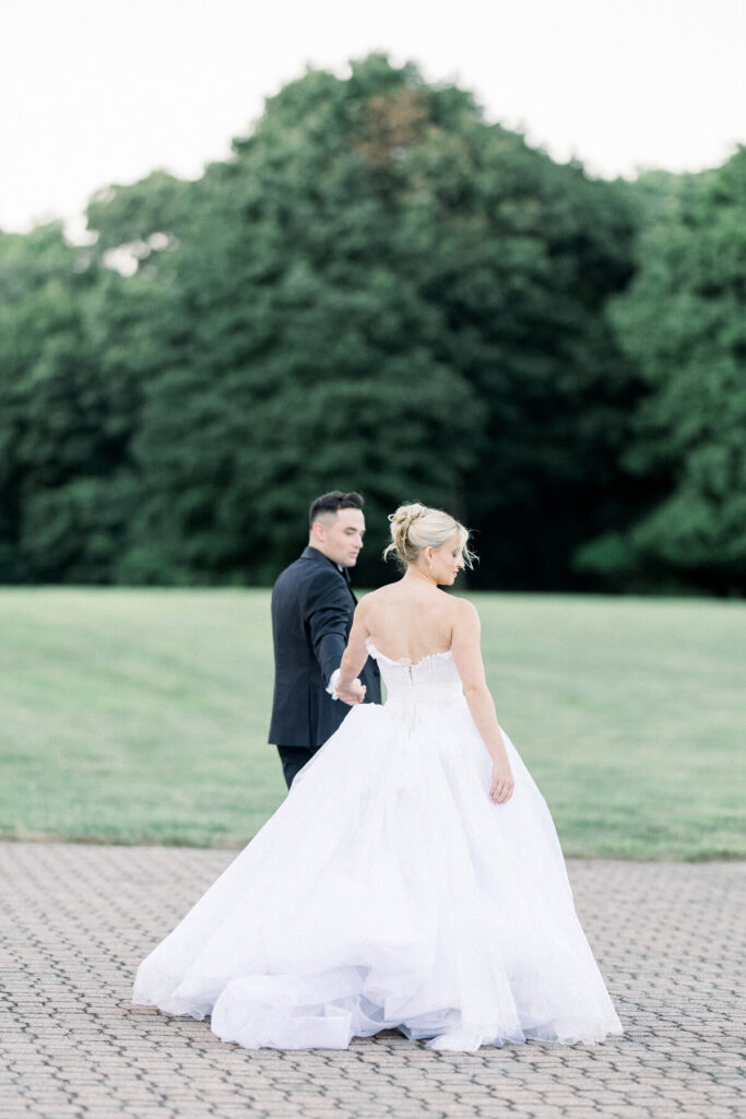 Newport Wedding Venues | Brianna Graca Weddings Fine Art Wedding Photographer in Newport | A couple in formal attire walks hand in hand, with the bride in a flowing white gown and the groom in a black suit, set against lush greenery. Romantic and serene atmosphere.