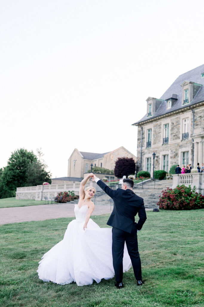 Newport Wedding Venues | Brianna Graca Weddings Fine Art Wedding Photographer in Newport | A bride in a flowing white gown and a groom in a black suit dance gracefully on a lush lawn, with an elegant stone mansion in the background, evoking joy and romance.